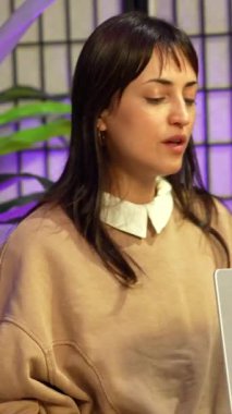 Genç iş kadını canlı yayına hazırlanıyor, laptopunu açıyor ve modern içeriğin oluşumunu yansıtan mor ortam aydınlatmasıyla çağdaş bir stüdyoda işine konsantre oluyor.