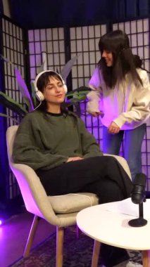 Bir diğer sunucu modern bir stüdyoda mor ışıklandırmalı bir dizüstü bilgisayar kullanırken, bir teknisyenin bir sunucuya lavalier mikrofonu ayarladığı bir podcast kaydına hazırlanan bir takımın sahne arkası görüntüsü