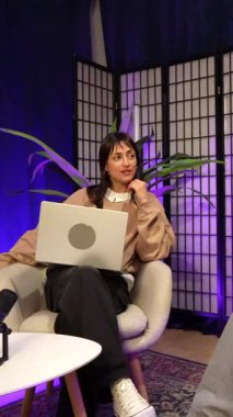 Genç erkek ve kadın koltuklarda oturuyor, podcast veya canlı yayın yaparken sohbet ediyorlar, çağdaş içerik oluşturma ve dijital iletişim kültürünü vurguluyorlar.