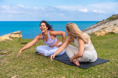 Yoga seansından zevk alan iki mutlu arkadaş ve mavi okyanusa bakan çimenli bir uçurumun kenarında gülüşmeler, bir anlık refah ve dostluk paylaşımı.