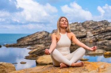 Yogadaki kadın deniz kenarındaki kayalık kıyı şeridinde poz veriyor, dinginlik, denge ve sağIık için idman yapıyor, dingin okyanus ufku ve doğal ışık arasında.