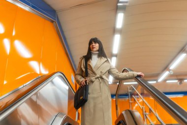 Metro istasyonunda yürüyen bir kadın, tırabzana tutunarak, trençkot giymiş, şehir hareketini, günlük rutinini ve toplu taşımayı yansıtıyor.