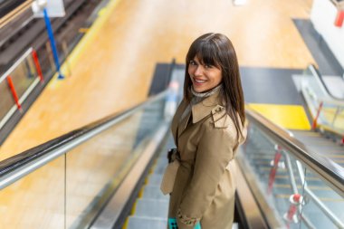 Trençkotlu bir kadın, modern bir metro istasyonunda yürüyen merdivenden inerken güvenle gülümsüyor. Şehir yaşam tarzını, günlük ulaşımı ve toplu taşımayı temsil ediyor.