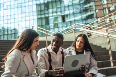 Diverse business professionals or students sitting on urban stairs, discussing project ideas and sharing information on a laptop during an informal outdoor meeting or study session