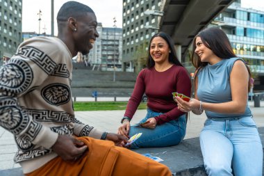 Çeşitli genç arkadaşlar birlikte gülerek dışarıda, modern bir şehir ortamında kart oynuyorlar, boş vakit ve eğlence anlarını paylaşıyorlar.