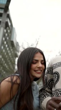 Mutlu, genç, çok kültürlü bir çift dışarıda birlikte cep telefonu kullanırken gülümsüyor ve gülüyor. Arka planda modern şehir binalarına yakın duruyor ve zamanlarının tadını çıkarıyorlar.