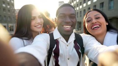 Şehirde selfie çekerken gülümseyen ve gülen üç genç arkadaştan oluşan mutlu çok ırklı bir grup. Arka planda güzel bir işaret fişeğiyle güneşli bir günün tadını çıkarıyorlar.