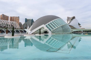 Hemisferic binası Valencia, İspanya 'daki sanat ve bilim şehrinde turkuaz gölde yansıyor. Etrafı fütürist, yenilikçi yapılar ve bulutlu gökyüzü ile çevrili.