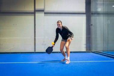 Gülümseyen, raket tutan bir bayan atlet ve sarı top canlı, mavi bir picleball sahasında, spor olsun diye spor yapan spor salonlarında spor yapan aktif boş zamanlarında.