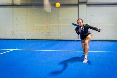 Kadın sporcu sarı bir picleball 'a raketiyle vuruyor. Kapalı alanda canlı mavi bir kortta. Yoğun bir oyun sırasında odak, çeviklik ve dinamik hareket gösteriyor.
