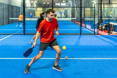 Arka planda diğer oyuncularla oynanan dostça bir maç sırasında sarı topa odaklanan parlak mavi bir penyball sahasında raket tutan bir adam.