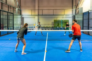 Dört farklı oyuncu, kapalı alanda oynanan mavi bir sahada, spor ve spor yaparken aktif olarak raket sallayarak, çiftler halinde maç yapıyorlar.