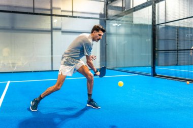 Mavi bir kapalı tenis sahasındaki erkek sporcu hızlı tempolu bir maç sırasında küreğiyle sarı bir topa vurmaya yoğun bir şekilde odaklanmış, çeviklik, hız ve kararlılık gösteriyor.