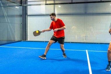 Kırmızı tişörtlü ve siyah şortlu bir adam aktif olarak canlı bir mavi sahada, bir oyun sırasında siyah bir kürekle sarı bir topa vuruyor.
