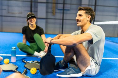 İki arkadaş, elinde raketlerle mavi bir Pickleball sahasında dinleniyor ve dinliyorlar, atletik aktiviteleri sırasında bir dakikalık dostluk ve stratejinin tadını çıkarıyorlar.