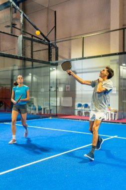 İki genç sporcu aktif olarak kapalı alanda canlı bir mavi sahada, dinamik ve iç açıcı bir maç sırasında yoğun bir şekilde sarı topa odaklanıyorlar.