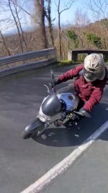 Motosiklet sürerken kask ve kırmızı ceket giyen bir insanın yüksek açılı bakış açısı. Doğayla çevrili, güneşli asfalt bir yolun kıvrımlarına ustaca eğilmiş.