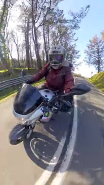 Kaskı ve ceketi olan bir kadın bisikletçi asfalt yolda motosiklet sürüyor, güneşli bir günübirlik gezinin tadını çıkarıyor parlak güneş ışığıyla dolu pitoresk bir orman manzarası