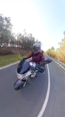 Kasklı ve ceketli bir kadının, güneşli bir günde, boş bir asfalt yolda, manzaralı bir virajda motosiklet sürerken görüntüsü.