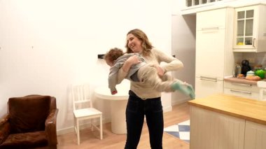 Neşeli beyaz anne, modern apartman mutfağında dans ederken kucağında gülen bebeğini döndürüyor. Saf mutluluk, sevgi ve aile eğlencesini paylaşıyor.