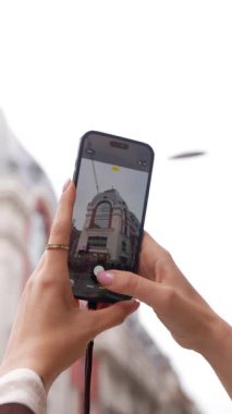 Şık bir şapka ve ceket giyen güzel bir kadın, akıllı telefonuyla eski bir binanın fotoğrafını çekiyor. Şehir sokaklarını geziyor ve beyaz arka planda tatilinin tadını çıkarıyor.