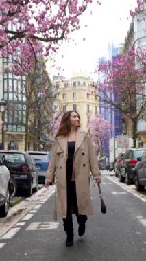 Trençkotlu güzel, mutlu bir kadın Paris 'in pitoresk bir caddesinde yürüyor, güzel bir bahar gününde çiçek açan pembe manolya ağaçlarının tadını çıkarıyor.