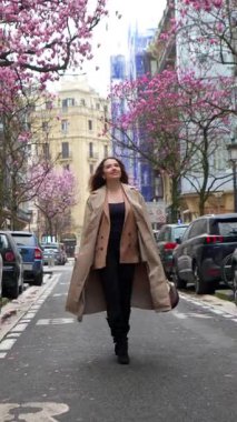 Trençkotlu güzel bir kadın Paris 'te bir caddede güvenle yürüyor. Etrafı güzel pembe manolya ağaçlarıyla çevrili ve bahar mevsiminin tadını çıkarıyor.