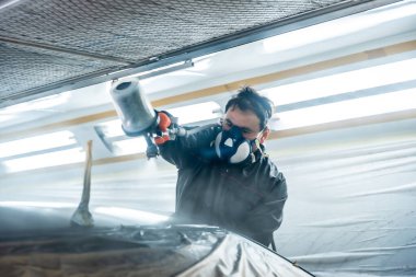 Otomobil teknisyeni koruyucu solunum cihazı takıyor ve araba parçalarını sprey silahla boyuyor, araç boyama kabininde profesyonel tamirat yapıyor.