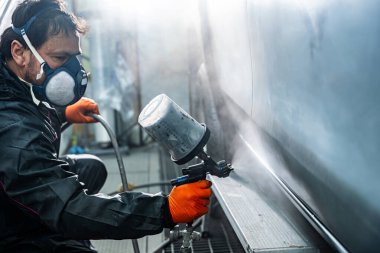 Solunum maskesi takan profesyonel oto boyacı ve koruyucu eldivenler ustalıkla bir atölyede sprey tabanca kullanarak araç bileşenine taze boya sürüyor.