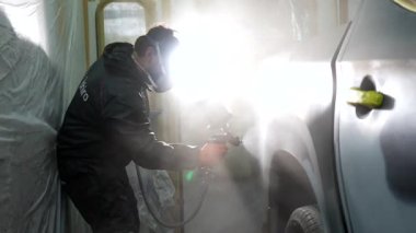 Profesyonel otomotiv ressamı solunum maskesi takıyor. Havalı fırça tabancası kullanıyor. Bir benzin istasyonundaki özel boya kabininde çalışan bir araca yeni bir kat boya sürüyor.