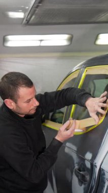 Kapıya ve pencereye dikkatlice maske bandı ve kağıt yapıştıran titiz bir kapıcı, bir tamirhanedeki profesyonel sprey kabininin içinde aracı boyamaya hazırlıyor.