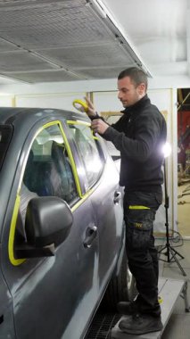 Profesyonel araba boyacısı, bir arabanın pencere çerçevesine titizlikle maske bandı yapıştırıyor, özel bir oto tamirhanesinde yeni bir boya işi için arabayı dikkatlice hazırlıyor.