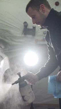 Profesyonel otomobil teknisyeni bir yağ çözeltisi püskürtüyor ve bir araba çamurluğunu mikrofiber bezle siliyor. Atölyede yeni bir boyama için araç yüzeyini titizlikle hazırlıyor.