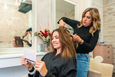 Modern bir güzellik salonunda müşterinin uzun kahverengi saçlarını düzelten profesyonel kuaför, saç bakımına ve dönüşüme odaklanıyor, müşteri gülümsüyor ve yansımasını kontrol ediyor.