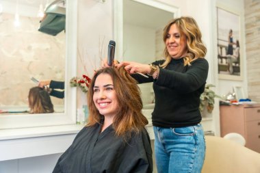 Kuaför, gülümseyen müşterinin uzun kahverengi saçlarını parlak bir güzellik salonunda düz ütüyle şekillendiriyor. Profesyonel bakım ve keyifli bir deneyim sağlıyor.