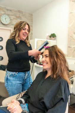 Profesyonel kuaför, güler yüzlü bir kadının saçını düz ütüyle düzeltiyor. Parlak, yoğun bir salonda, dostane bir müşteri randevusu sırasında cilalı yeni bir görünüm yaratıyor.