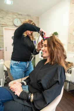 Kuaför, saç maşası kullanan, saç maşası kullanan, modern güzellik salonu ortamında yeni bir saç stili oluşturan, profesyonel hizmet ve kişisel bakımı vurgulayan bir bayan müşteri üzerinde çalışıyor.