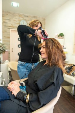 Kadın kuaför, gülümseyen bir müşterinin kahverengi saçlarını modern bir salonda ısıtmalı bir aletle şekillendiriyor. Şımartma, güven ve uzman saç bakım hizmetleri sunuyor.