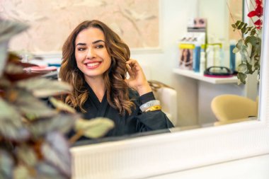 Dalgalı kahverengi saçlı genç bir kadın aynadaki yansımasına gülümsüyor, yeni bir saç kesiminin tadını çıkarıyor ve profesyonel bir davranışın ardından kendini şımartıyor.