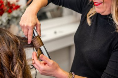 Profesyonel kuaför, kuaförde bir kadının uzun esmer saçlarını saç maşasıyla dikkatlice şekillendiriyor. Güzellik ve şık bir görünüm için bukleler yapıyor.