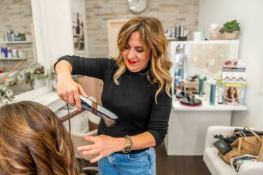 Profesyonel kuaför, müşterinin uzun, kahverengi saçlarını ütüyle dikkatlice şekillendiriyor dalgalar yaratıyor ve modern bir kuaför salonunda güzellik hizmeti veriyor.