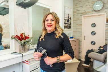 Kuaföründe elinde makas, tarak ve fırça ile gülümseyen, kendinden emin ve misafirperver, uzmanlık ve dostça hizmet sunan müşterileri stile sokmaya hazır profesyonel bir bayan kuaför.