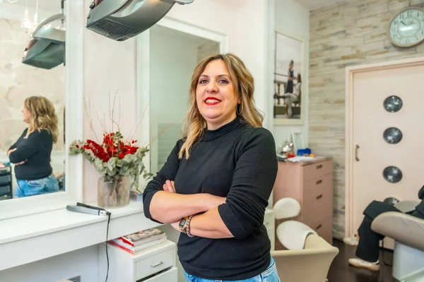 Kolları çapraz duran kadın kuaför, kuaföründe kameraya gülümsüyor, küçük işletme sahipliğini ve profesyonel güzellik hizmetlerini vurguluyor.