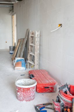 Kirli bir kırmızı kova, alet çantası, merdiven, inşaat malzemeleri ve borularla yenileme işlemini gösteren boş oda, devam eden bir bina veya kurulum projesini işaret ediyor