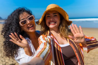 Mutlu kadın arkadaşlar yaz tatilinin tadını çıkarıyorlar. Kameraya gülümsüyor ve el sallıyorlar. Bir yandan da selfie çekiyorlar.
