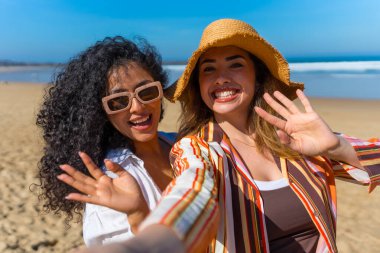 Güneşli bir plaj tatilinde olan iki neşeli genç kadın neşeli bir selfie çekip, güneş şapkaları ve güneş gözlükleriyle kameraya el sallıyorlar, rahat ve tasasız bir şekilde.