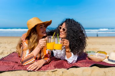 İki neşeli bayan arkadaş kumlu bir plajda yaz pikniğinin tadını çıkarıyorlar, kadeh kaldırarak bir kadeh portakal suyu kaldırıyorlar, açık mavi gökyüzü altında tatil ve dostluğu kutluyorlar.