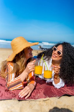 İki neşeli genç kadın kumda bir battaniyeye uzanmış, bir bardak taze portakal suyuyla kadeh kaldırıyor, arkadaşlıklarını kutluyor ve okyanusun kenarında rahatlatıcı bir yaz gününün tadını çıkarıyorlar.