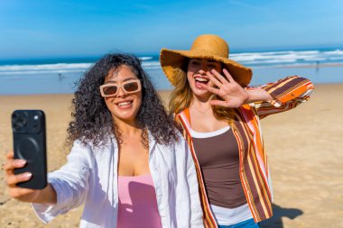 Güneşli kumlu bir sahilde selfie çeken iki neşeli genç kadın okyanus dalgalarının yanında gülüyor ve poz verirken kaygısız bir yaz tatili geçiriyorlar.