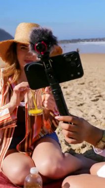İki kadın içerik yaratıcısı güneşli bir plajda, piknik yaparken ve deniz kenarında yaz tatillerinin tadını çıkarırken mikrofonla akıllı telefon kullanarak bir seyahat videosu çekiyorlar.